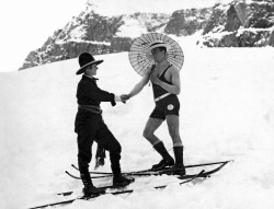 Adam Glickman - A woman skier on the mountain slope wearing a sash and western hat shakes hands with a man on skis in a one piece bathing suit and carrying a parasol, early to mid 1920&rsquo;s.