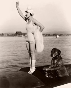 Gloria Swanson (1899 - 1983) stands on tiptoes on the prow of a motorboat while Teddy the dog sits with his paws on the steering wheel in a still from director Clarence G Badger&rsquo;s film Teddy at the Throttle, 1916.