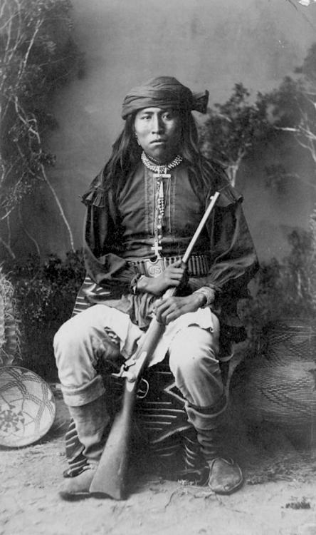 thebigkelu:  Portrait of Nosey San Carlos Apache Scout, in Native Dress with Gunbelt and Gun; Blanket and Baskets Nearby - Randall - 1886