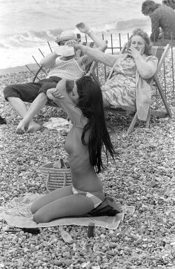 historium:Woman covering man’s eyes with her knitting at sight of young woman taking off her top on the beach in France, 1974