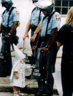 unexplained-events:  This photo was taken over 20 years ago by Todd Robertson during a KKK rally in northeast Georgia. One of the boys approached a black state trooper, who was holding his riot shield on the ground. Seeing his reflection, the boy reached
