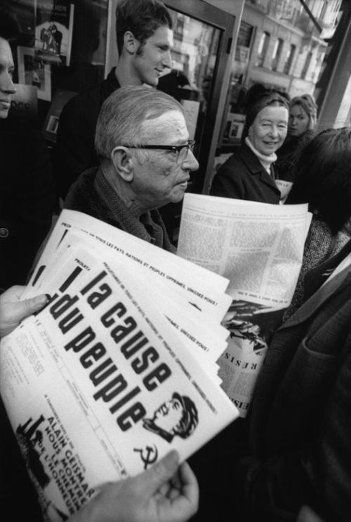 Jean-Paul Sartre avec Simone de Beauvoir (au fond, le regardant en souriant) distribuant le journal maoïste La Cause du Peuple, interdit par le gouvernement français, à Paris en 1970. Nudes &amp; Noises  