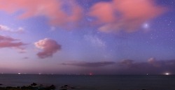The Earth’s clouds reflecting moonlight, creating a faint, reddish glow at a beach in northern France. Beyond the clouds lie cosmic dust and star clouds of the Milky Way. The constellation Sagittarius can be seen peaking above the horizon and